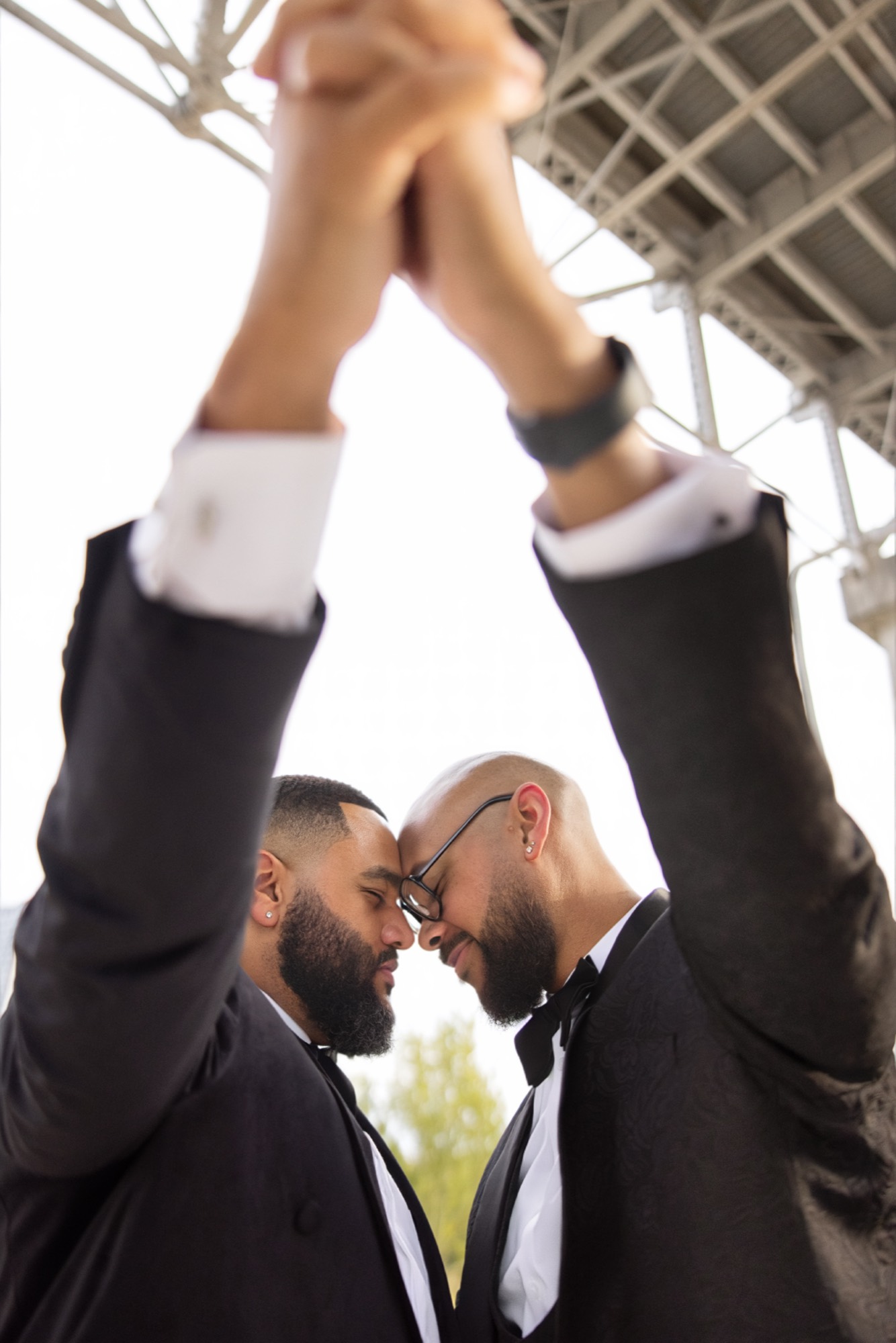 Aaron and Deonte in tuxedos, foreheads touching, hands raised together beneath a steel bridge